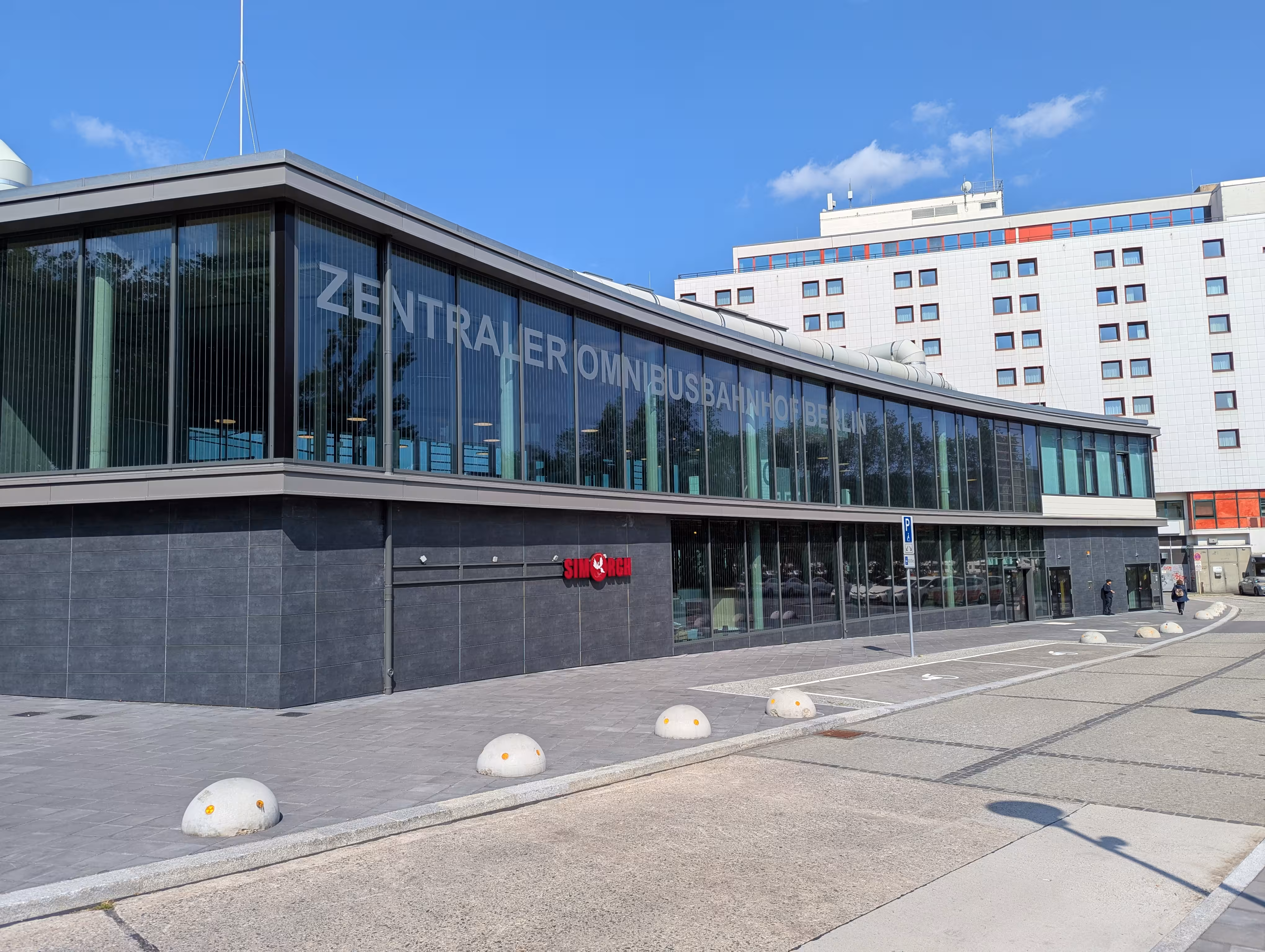 Die Wartehalle am ZOB von außen mit zwei Behindertenparkplätzen davor und dem ibis Hotel im Hintergrund